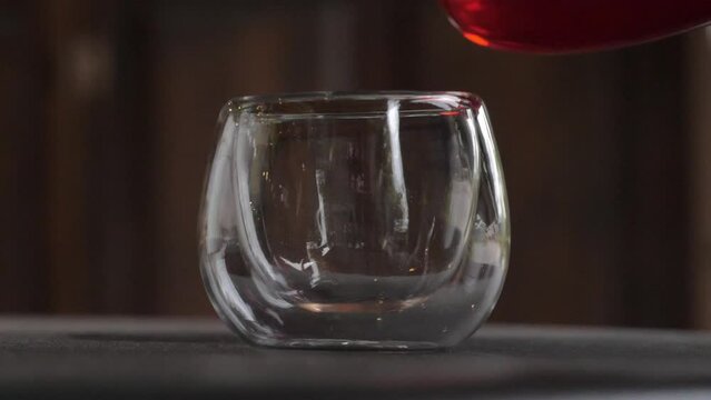 Slow motion speed closeup scene on stream of dark orange color ceylon tea pouring from teapot to fill in empty glass isolated on black background, beautiful style transparent teacup.