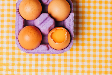 four chicken eggs in purple egg box on yellow plaid tablecloth, top view
