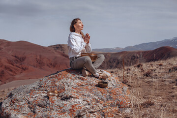 Naklejka na ściany i meble Meditation in rocky mountains. Girl sits on a stone with folded hands in a lotus position with her eyes closed. There is a singing bowl at her feet.
