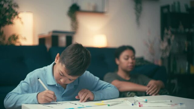 Little boy drawing picture home, depressed mother sitting behind, divorce crisis