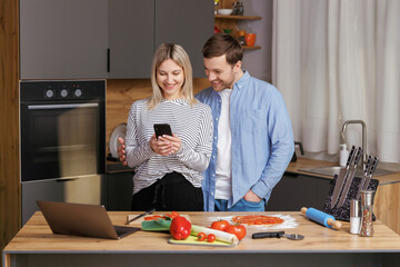 Smiling loving couple cooking salad together while standing on a kitchen at home and using mobile phone. Cute couple man and woman taking photo of food and taking pictures together while cooking.