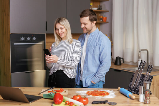 Smiling loving couple cooking salad together while standing on a kitchen at home and using mobile phone. Cute couple man and woman taking photo of food and taking pictures together while cooking.