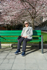 Fototapeta premium an eighty-year-old woman sunbathes in sunglasses sitting on a green wooden bench under a Japanese cherry tree