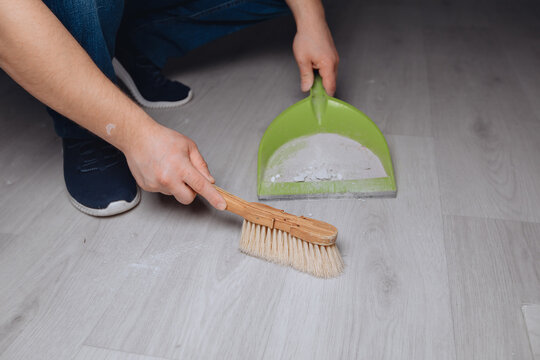 A young man is squatting and sweeping the dust with a brush after repair work in a dustpan. Completion of construction. - Powered by Adobe
