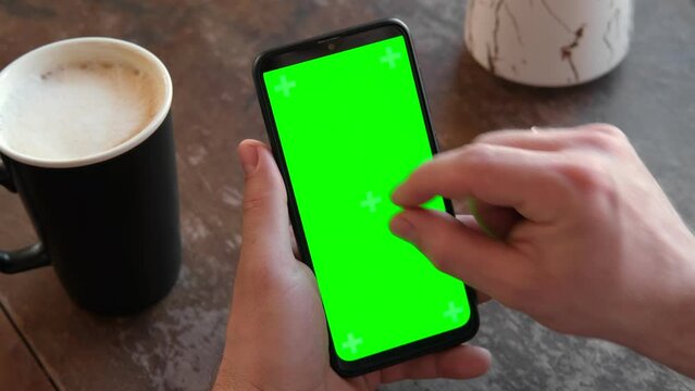 Man Use of Mobile Phone with Blank Green Screen in a Cafe on Rustic Vintage Wooden Table. Male Holding Smart Phone Empty ChromaKey Screen Mock up in Coffee Shop. Morning. Showing Mock up Video Footage