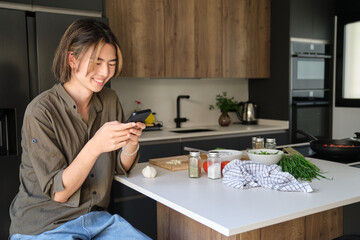 Smiling asian young man checking recipe on mobile phone at kitchen.