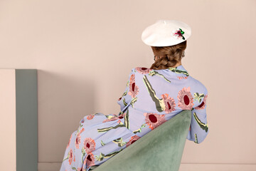 Rear view of unrecognizable woman in hat sitting in chair on background of beige wall, fashion shoot