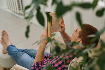 Man leisurely reading a book in a comfortable chair, nestled within verdant foliage indoors....
