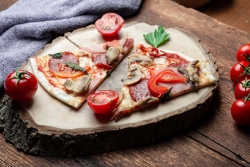 Pizza with ham and bacon, mushrooms and tomatoes on a slice of wood on a brown wooden background.