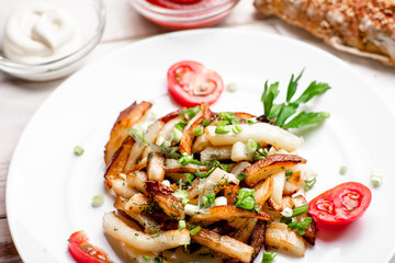 Fried potatoes in a white plate with green onions, parsley, tomatoes on a white wooden background.