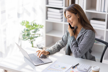 Attractive successful elderly business Asian woman in striped blouse working in modern office, making phone call to potential client, having nice conversation, sitting at desk in front of open laptop
