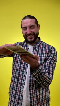 Happy mid adult bearded man throwing bunch money into camera scattering banknotes, rejoicing luck lottery, boasting money profit. Indoor vertical shot isolated yellow background. Money to burn concept