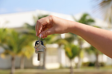 Real estate agent, home keys in female hand on background of house surrounded by palm trees. Buying or rental a villa on ocean coast, removal to tropical country