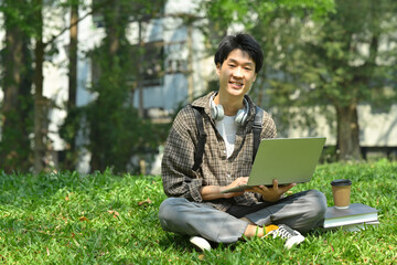 Smiling asian man student using laptop on green grass in front of university building. Education, technology and lifestyle concept