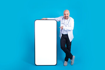 Big smartphone, cheerful smiling middle aged doctor pointing big smartphone. Bald physician leaning huge mobile phone with empty screen, mock up. Isolated blue studio background, copy space. 