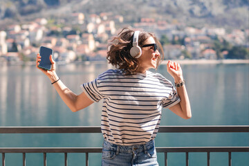 Young happy woman listening to music using headphones while standing on the balcony with amazing lake and mountain views