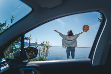 Happy cheerful young woman traveling on the sea by car. Standing overlooking the sea with hands up - 586517532
