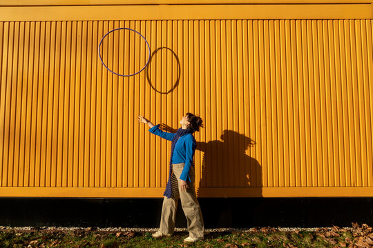 Young woman tossing hula hoop near yellow container on sunny day