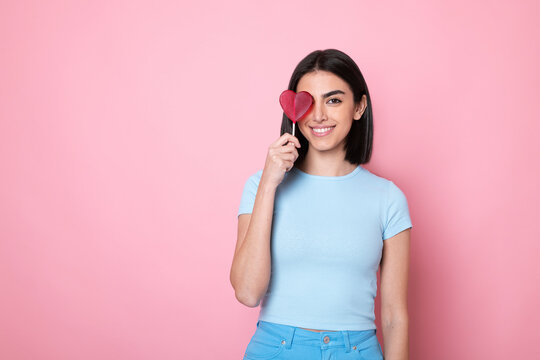 Happy woman holding heart shaped lollipop over eye against pink background