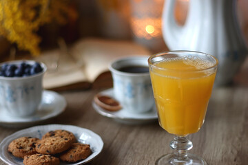 Cup of tea or coffee, plate of cookies, cup of blueberries, plate of chocolate, glass of juice, book, reading glasses, teapot, flowers and lit candles on the table. Brunch or afternoon tea concept.