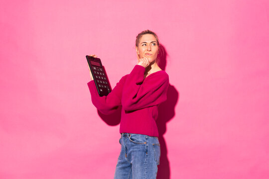 Confused woman with calculator standing against pink background