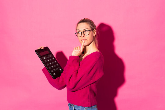 Thoughtful woman gesturing and holding calculator against pink background