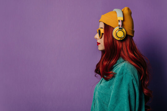 Redhead woman listening to music through headphones against purple background