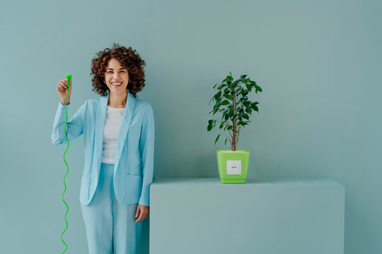 Happy woman with electric plug standing by potted plant in front of blue wall