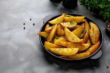 Roasted potatoes with herbs and spices. Baked potato wedges in frying pan on dark stone background. Copy space. - obrazy, fototapety, plakaty