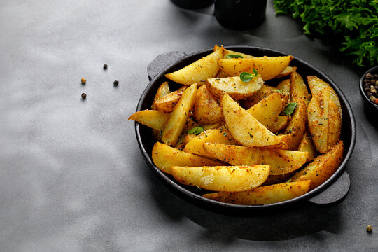 Roasted potatoes with herbs and spices. Baked potato wedges in frying pan on dark stone background. Copy space.