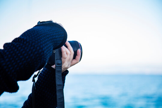 Hands Of Person With Photo Camera Lens Is Taking Photos On Sea Coast Against Blue Water Background, Copy Space, Space For Text 