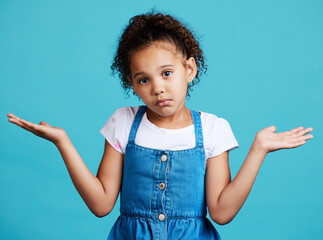Portrait, children and shrug with a girl on a blue background in studio feeling confused or lost. Kids, question and doubt with a young female child shrugging her shoulders to gesture whatever