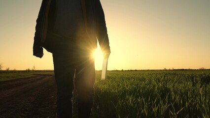 Farm work with digital tablet in agriculture. Farmer walks in boots with a computer tablet in green field of wheat sprouts at sunset. Technology of modern agriculture. Concept of environmentally