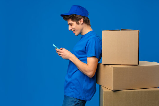 Young courier man in blue uniform smiling look at his smartphone while leaning on cardboard box parcels