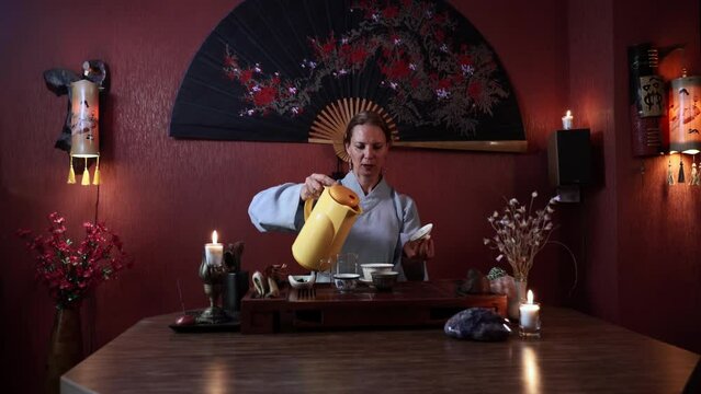 brewing pu erh tea, an adult woman pours hot water into a teapot during a tea ceremony while sitting at a table indoors