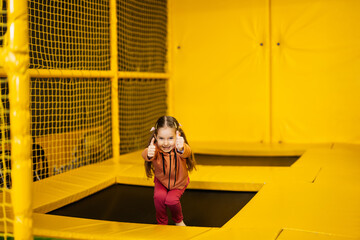 Fototapeta na wymiar Little girl kid show thumb up on trampoline at yellow playground park. Child in active entertaiments.