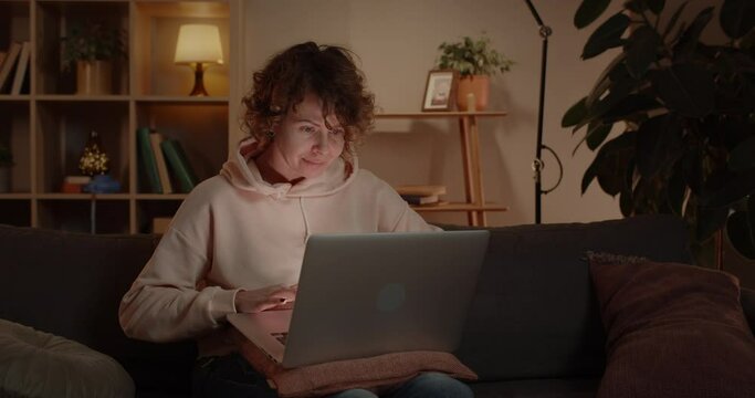 Cheerful woman using laptop while sitting on sofa at home. Female joyful person browsing internet and working with computer in living room. People and technology concept.