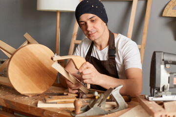 Portrait of focused handsome young carpenter wearing apron and cap working in workshop, holding...