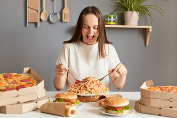 Portrait if amazed excited extremely hungry woman wearing white T-shirt sitting at table in kitchen, delivering lots jink food dishes, eating noodles, expressing happiness.