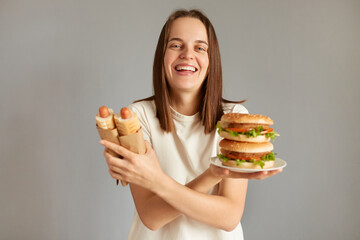 Portrait of laughing woman holding tasty cheeseburger and hotdogs, crossed hands, looking at camera with cheerful happy expression, likes unhealthy food.
