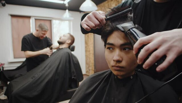 Professional barber slowly and carefully dries an asian man's hair with a hair dryer and a hairbrush close up. Man getting his hair done at a barbershop