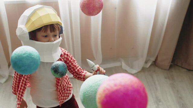 Caucasian child with a toy astronaut helmet on his head plays with a toy rocket between hanging toy planets standing at home in a children's room with curtains in the background