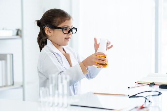 In Chemistry Classroom With Many Laboratory Tools On Shelves And Table. A Little Asian Girl With Glasses In White Lab Coat Holding Yellow Chemical Flask In Right Hand With White Stained In Left Hand.