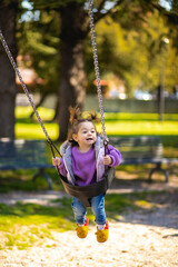 Cute caucasian little toddler girl with two tails smiling widely and swinging on a baby swing in the playground with violet sweeter sleeveless jacket jeans
