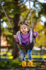 Sweet caucasian little toddler girl with two tails smiling widely and swinging on a baby swing in the playground with violet sweeter sleeveless jacket jeans