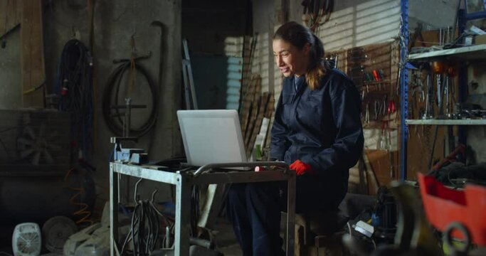 Portrait of Young Female Mechanic in Uniform Overalls Using Laptop Computer to Have a Video Call Chat with Family and Friends. Professional Woman Working as a Welder Taking a Break in her Workshop