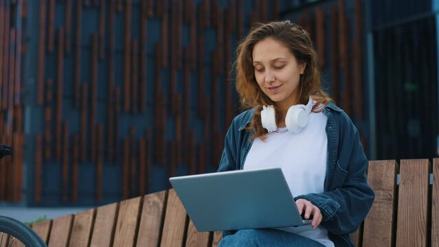 Caucasian smiled woman freelancer sitting on the bench working on the laptop near the office centre. Business Woman enjoy outside entrepreneur browsing typing. People and technology concept.