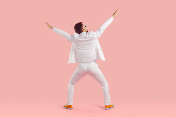 Fototapeta na wymiar Funny inspired man in white suit performs energetic fashionable dance stands with back to camera stretching arms up and hangs out having carefree time stands on studio pink background