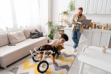 Smiling man using laptop near african american girlfriend and handicapped dog at home.