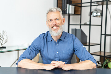 Headshot of handsome good looking mature man sitting at the office desk and looking at the camera with cheerful smile. Portrait of charismatic grey-haired senior man